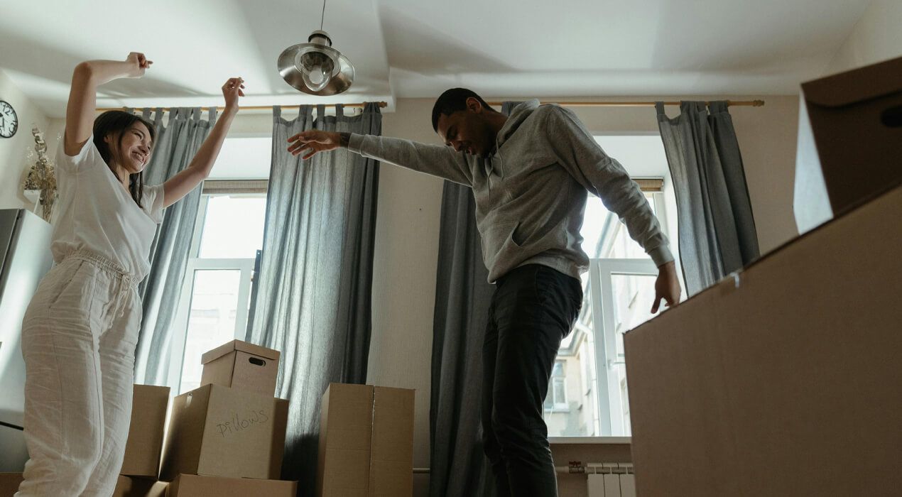 Young couple dancing around their living room, surrounded by moving boxes