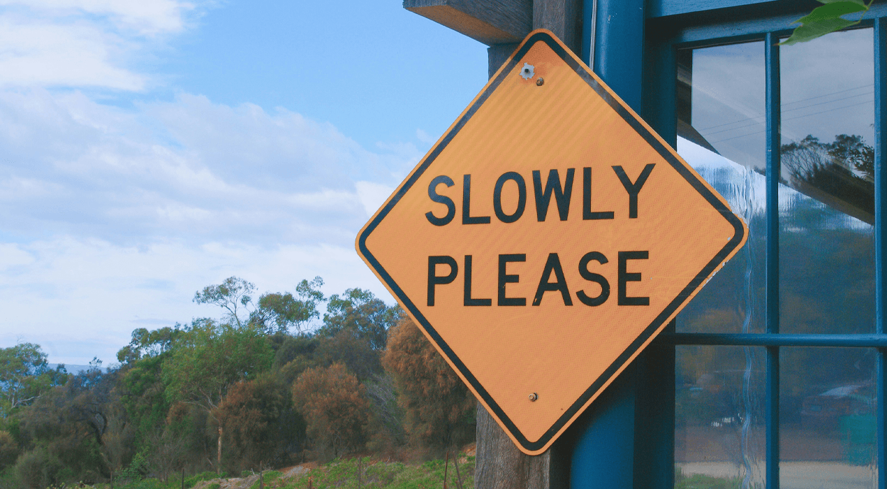 Yellow road sign reading “Slowly Please” hanging outside a building, with trees and blue sky in the background.