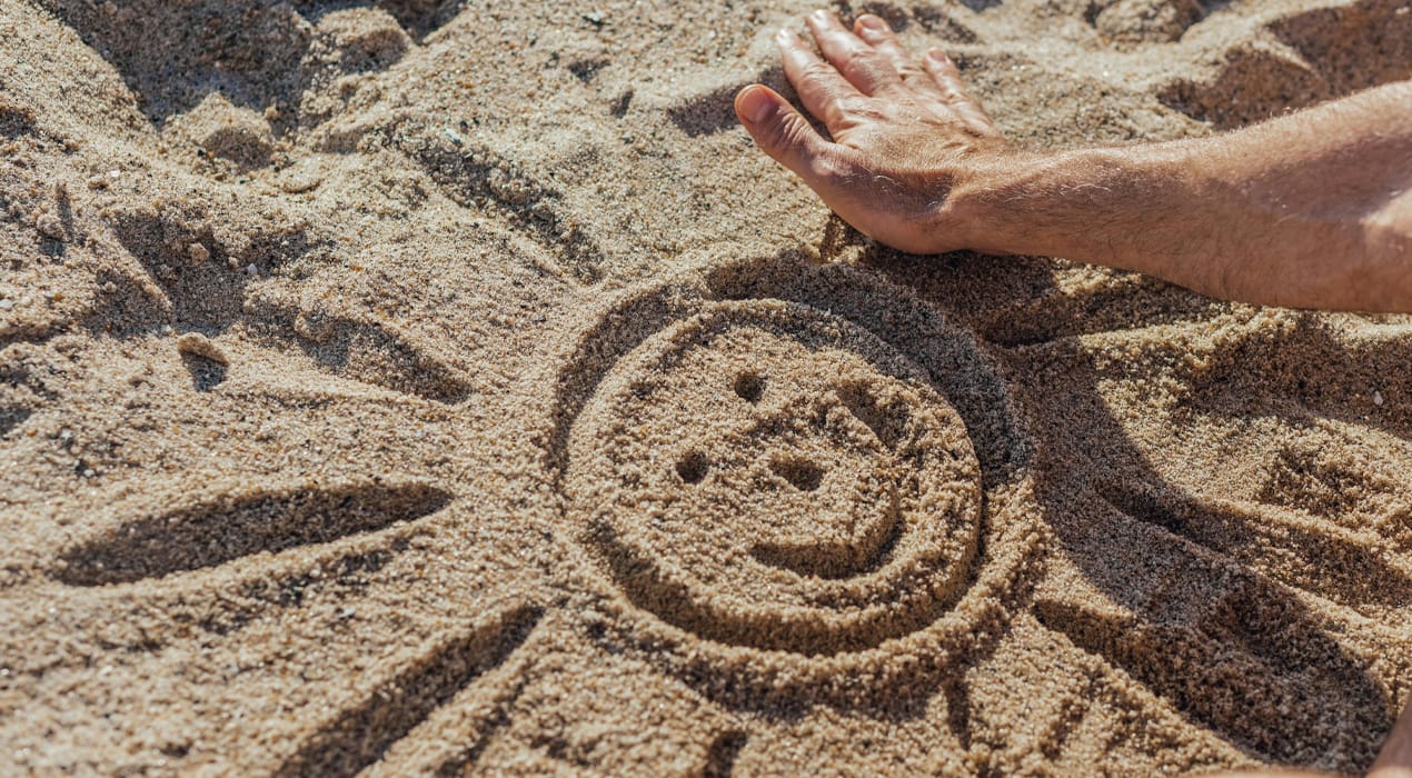 Person drawing a smiley face sun in sand