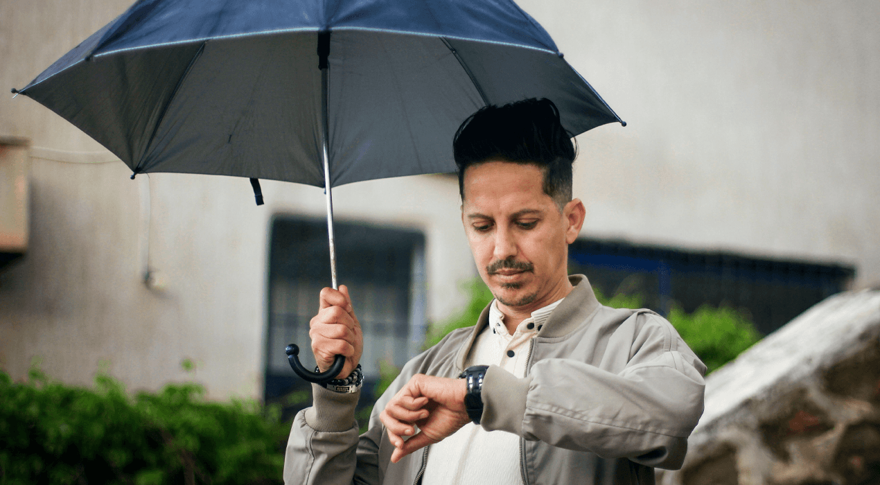 Man standing holding a black umbrella above his head and looking at his watch.