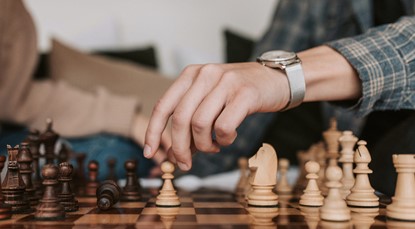 Pictured is a man's hand and forearm, about to move a white pawn on a chess board. The man wears a silver wristwatch and a blue and cream plaid shirt.