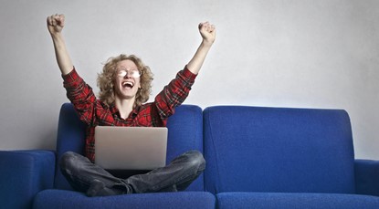 Young person sitting cross legged on a blue couch, with a laptop in their laptop, cheering with both arms up in the air