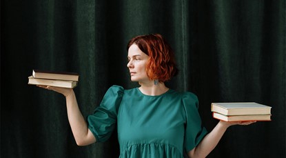 Woman standing against a green curtain, balancing two books in the palm of each hand—looking like she's weighing them up