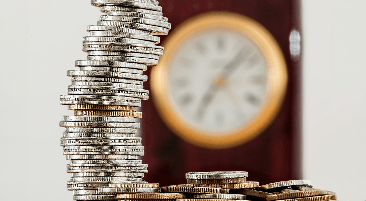 Big stack of coins and small pile of coins, with a clock in the distant background